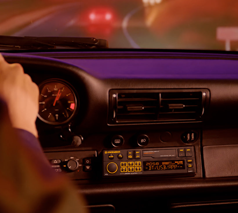 A close-up view from inside a car showing a hand on the steering wheel, the dashboard, instrument gauges, air vents, and a retro-style cassette radio with illuminated yellow buttons and display.