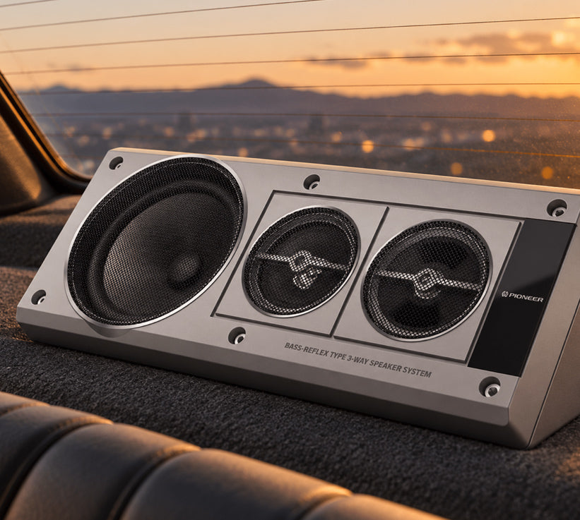 A silver and black Pioneer bass reflex speaker system installed inside a car, with a warm sunset cityscape visible through the window behind it.