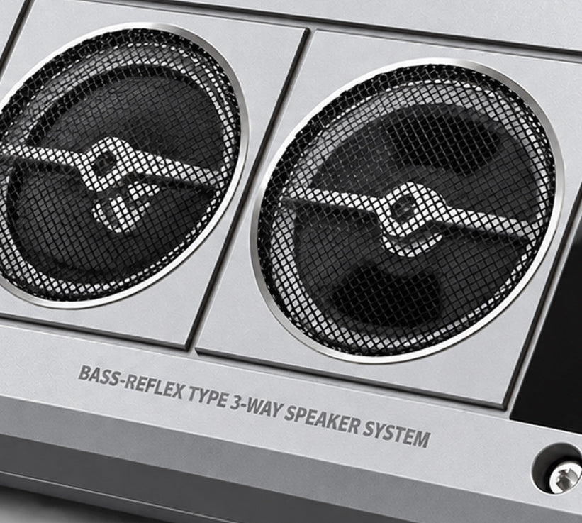 Close-up of a silver 3-way bass-reflex speaker system with two large black mesh-covered drivers and printed text reading 'BASS-REFLEX TYPE 3-WAY SPEAKER SYSTEM'.